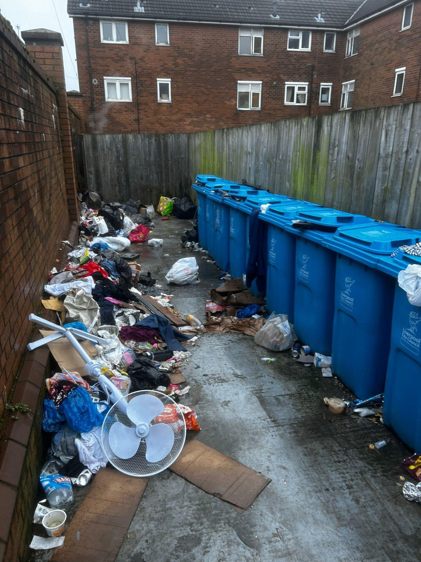 Long line of fly tipped clothing, bags and bulky items dumped beside Liverpool City Council recycling bins